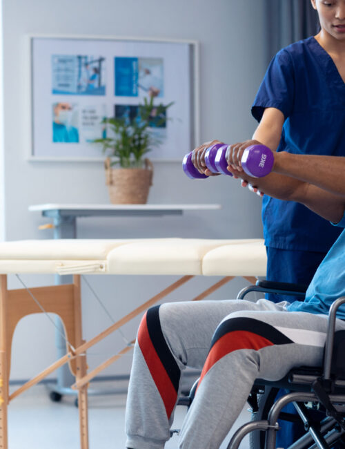 Diverse female physiotherapist helping senior male patient in wheelchair using dumbbells, copy space. Hospital, medical and healthcare services.